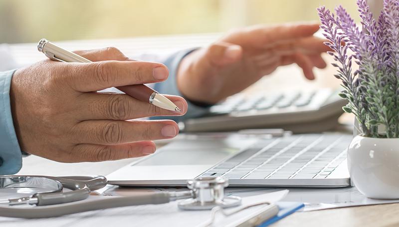 Doctor working on laptop computer with report analysis and money about Healthcare costs and fees in medical hostpital office. Focus stethoscope on table. Healthcare budget and business concept